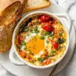 Feta Eggs – Easy Baked Breakfast Packed with Flavor 12 Baked feta eggs with cherry tomatoes, spinach, and herbs in a white ramekin, served with crusty bread on a plate.