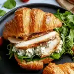 Golden Chicken Caesar Croissant Sandwich with grilled chicken and leafy greens on a dark plate.