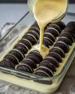 Pouring creamy batter over stacked chocolate sandwich cookies in a baking dish for Oreo dump cake