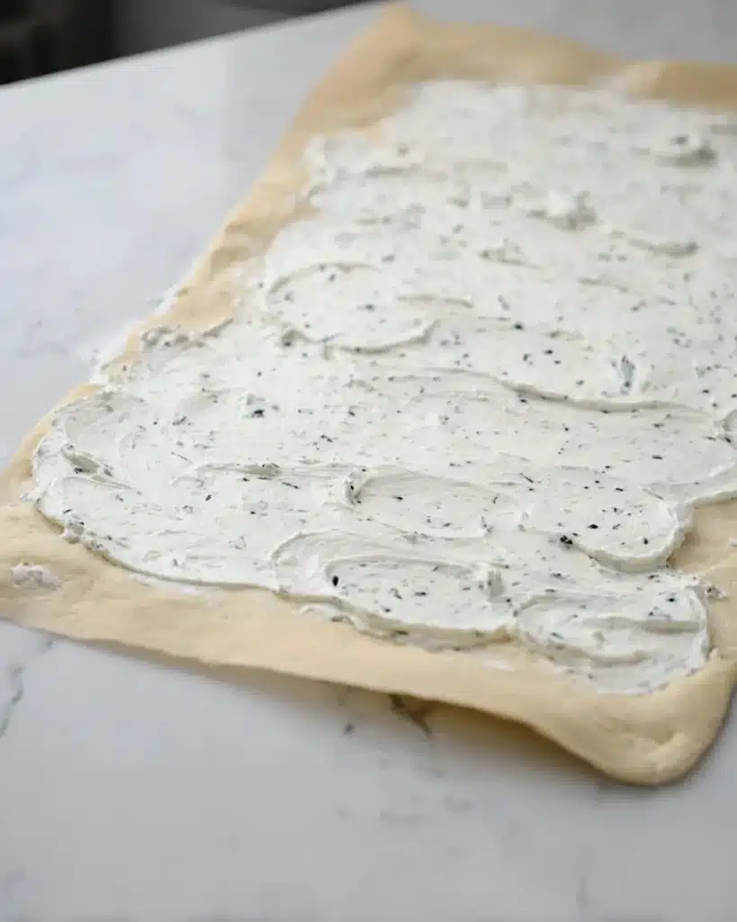 Cream cheese spread over crescent dough for breakfast roll preparation on a marble surface