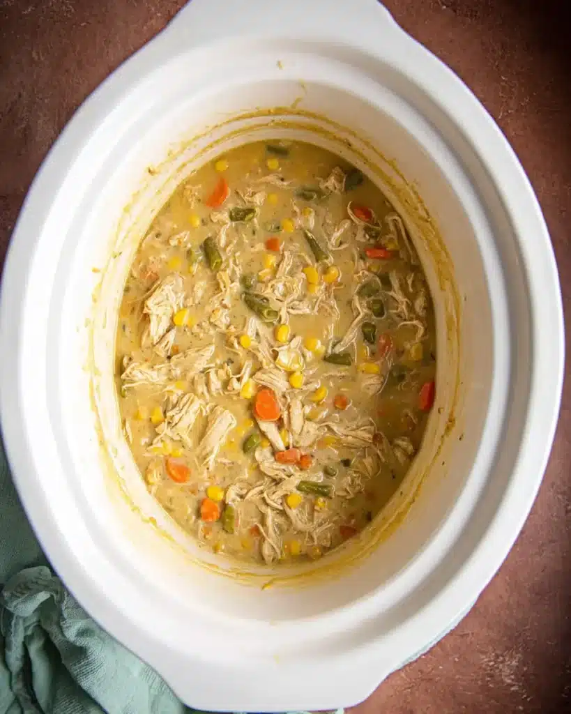 Cooked chicken pot pie filling with shredded chicken and vegetables in a white crockpot.