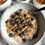 Bowl of creamy overnight oats topped with blueberries, granola, and peanut butter drizzle on a rustic table.