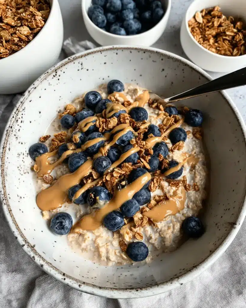 Bowl of creamy overnight oats topped with blueberries, granola, and peanut butter drizzle on a rustic table.
