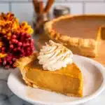Pumpkin Pie with Sweetened Condensed Milk | Remarkable Creamy Favorite 12 Slice of pumpkin pie with cinnamon whipped cream on white plate, fall-themed decor in background