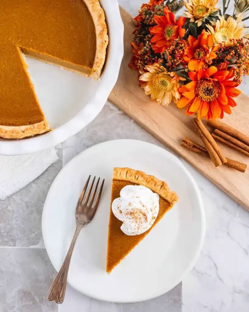 Pumpkin Pie with Sweetened Condensed Milk | Remarkable Creamy Favorite 10 Top-down view of pumpkin pie slice with cinnamon whipped cream on white plate, autumn decor and full pie in background