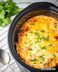 Slow cooker chicken enchilada casserole topped with melted cheese and herbs in a black Crockpot with spoon and cilantro on the side