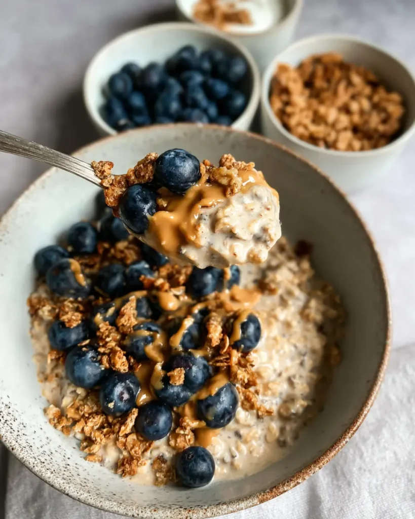 Spoonful of creamy overnight oats with blueberries, granola, and nut butter lifted above a breakfast bowl.

