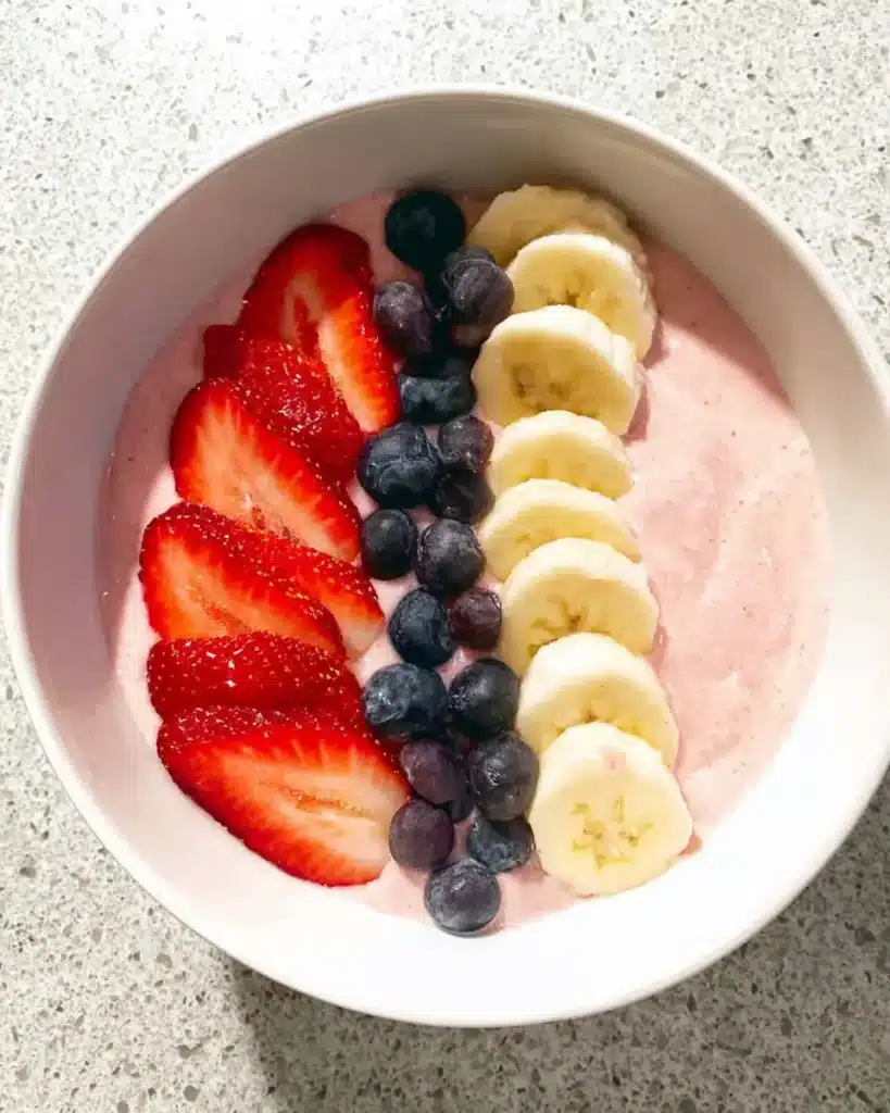 Colorful fruit smoothie bowl with pink strawberry base, topped with banana slices, strawberries, and blueberries