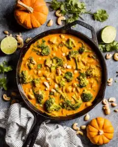 Colorful Thai pumpkin curry in a cast iron skillet with broccoli, cashews, and fall garnishes on a rustic table