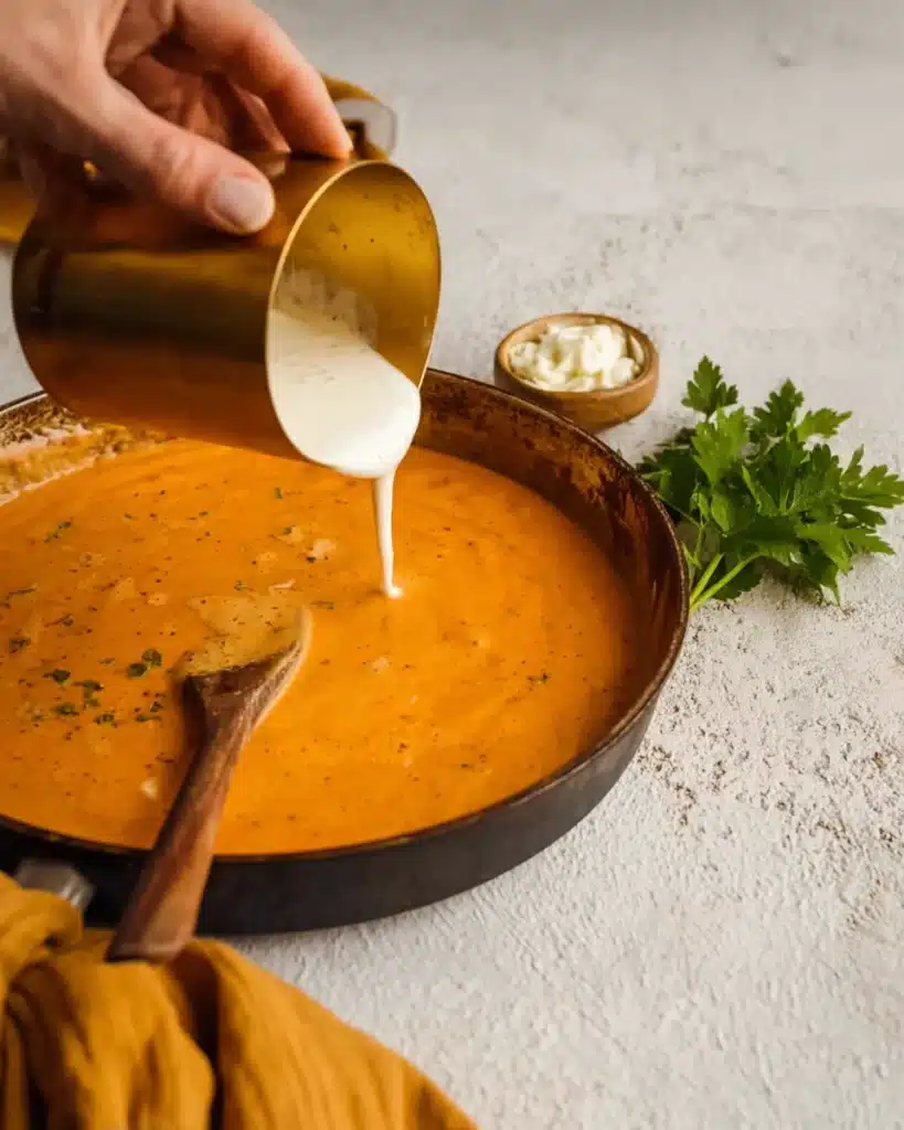 Pouring sour cream into pumpkin sauce for creamy pasta in a skillet with a wooden spoon and fresh parsley nearby.