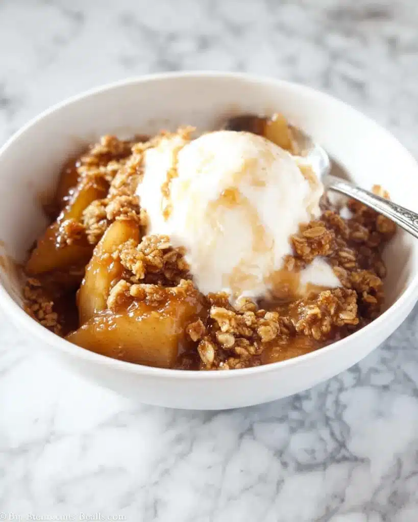 Warm apple crisp topped with vanilla ice cream in a white bowl on marble surface