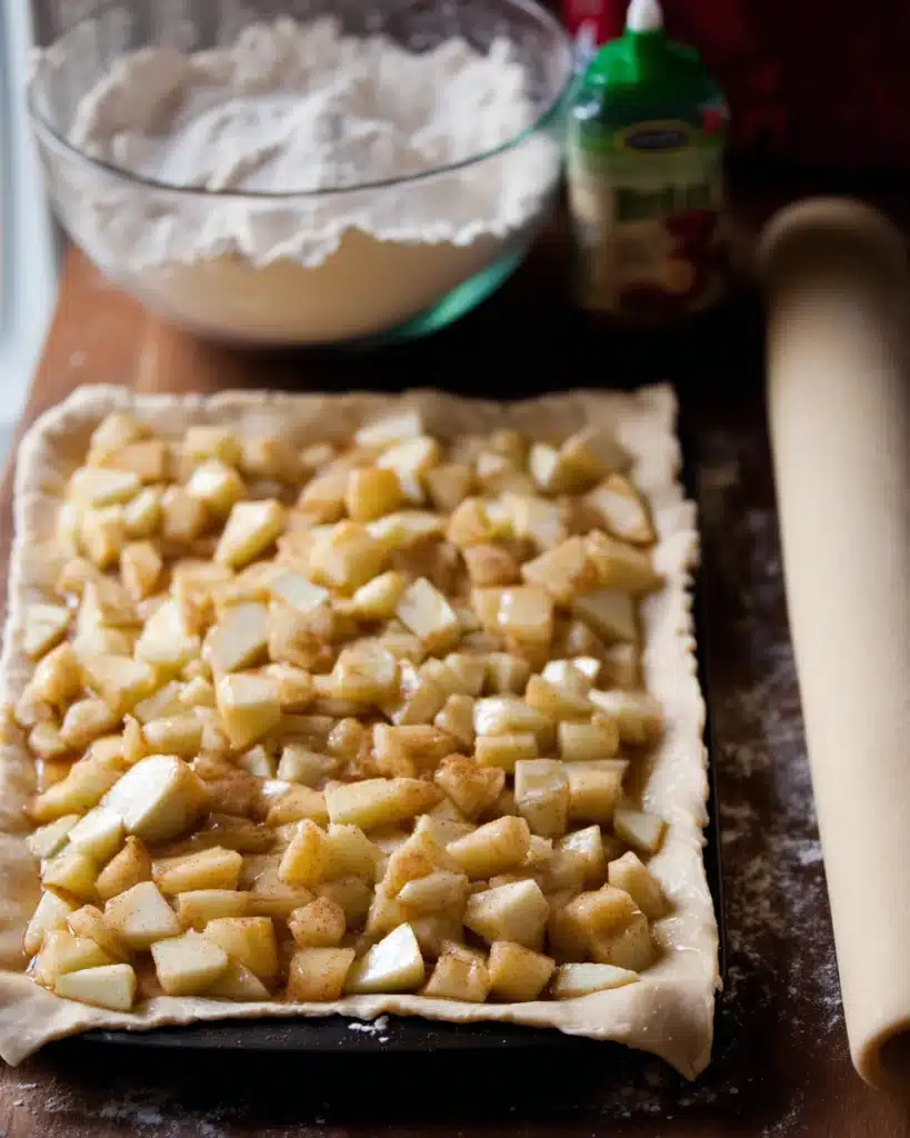 Raw apple slab pie being assembled in a pan, filled with diced apples over soft pie crust, ready for baking.