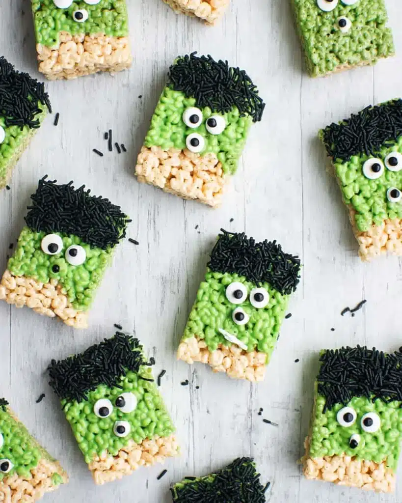 Multiple Halloween Rice Krispie treats decorated as green Frankenstein monsters with candy eyes and black sprinkles on a light tabletop.