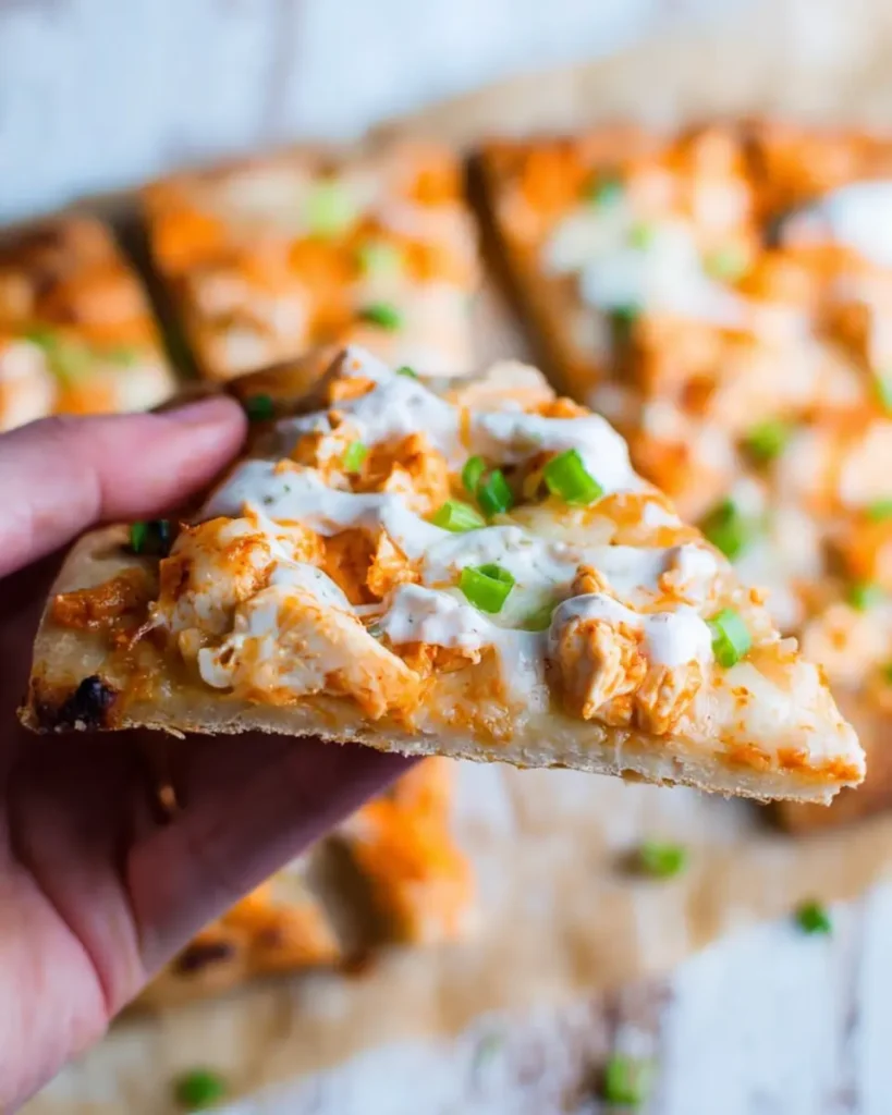 Hand holding a slice of buffalo chicken flatbread topped with cheese, buffalo sauce, chicken, and green onions.