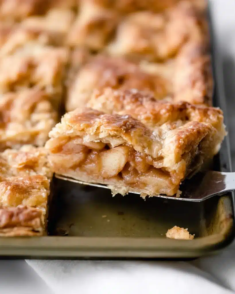 Golden apple slab pie slice lifted from the pan, showing layers of flaky crust and gooey spiced apple filling in close-up detail.