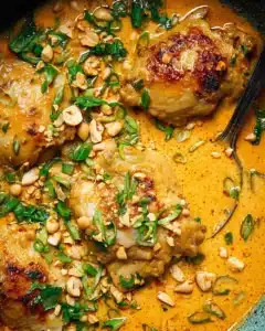 Close-up of creamy coconut lime chicken thighs topped with chopped peanuts, scallions, and cilantro in a rich orange sauce
