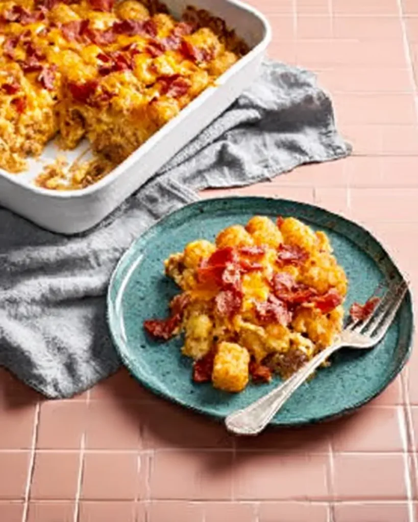 Tater Tot Sausage Breakfast Casserole with crispy bacon, served on a teal plate beside the full casserole dish on pink tile.

