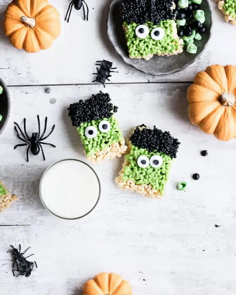 Top-down view of Halloween Rice Krispie treats decorated as green monsters with candy eyes, black sprinkles, pumpkins, spiders, and a glass of milk.
