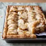 Freshly baked apple slab pie in a rectangular pan with golden, flaky crust and juicy apple filling, served on a towel with pie slice being lifted.
