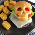 Halloween cheeseburger skull with golden bun, melted cheese patty, lettuce, tomato, and crispy tater tots on a plate