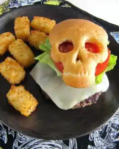 Halloween cheeseburger skull with golden bun, melted cheese patty, lettuce, tomato, and crispy tater tots on a plate