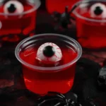 Red jello shots with floating candy eyeballs in clear cups on a black lace background, decorated with a plastic spider for Halloween.