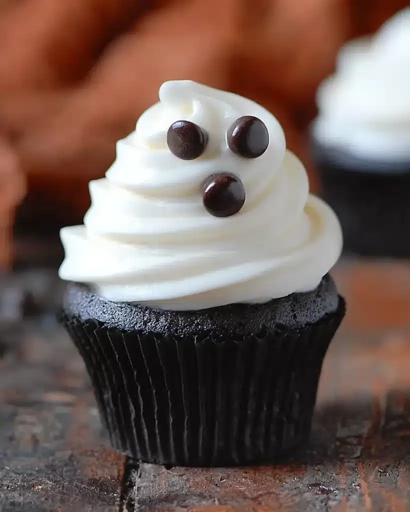 Close-up of a Halloween ghost cupcake with white frosting swirls and chocolate chip eyes and mouth.