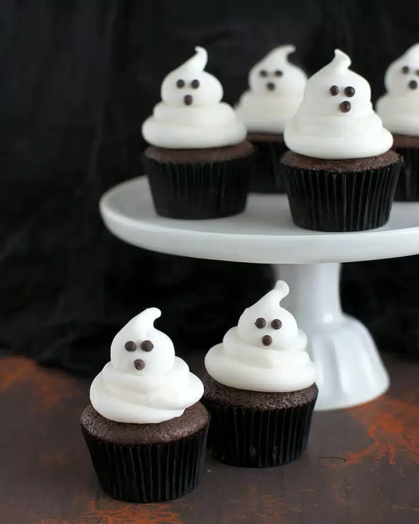 Chocolate cupcakes on a white stand decorated with ghost-shaped frosting and chocolate eyes, perfect for Halloween parties.