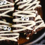 Halloween mummy brownies with white chocolate drizzle and candy eyeballs on a dark tray