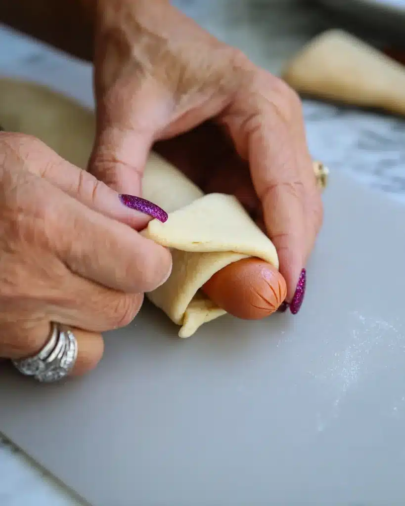 Hands wrapping crescent roll dough around a hot dog for spooky spider hot dogs.