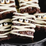 Mummy Brownies – Easy Halloween Treats 14 Halloween mummy brownies with white chocolate drizzle and candy eyeball decorations on a rustic table.