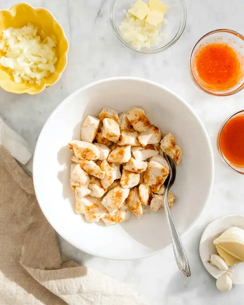 Seared chicken cubes tossed in ranch dressing with buffalo sauce, garlic, butter, and onion prepped in bowls around a white plate.