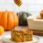 Pumpkin baked oatmeal slice topped with honey drizzle on a white plate, with a honey dipper above and a blurred pumpkin in the background.