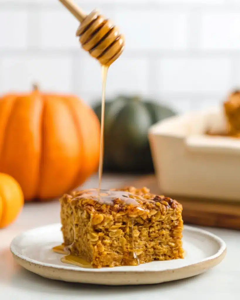 Pumpkin baked oatmeal slice topped with honey drizzle on a white plate, with a honey dipper above and a blurred pumpkin in the background.