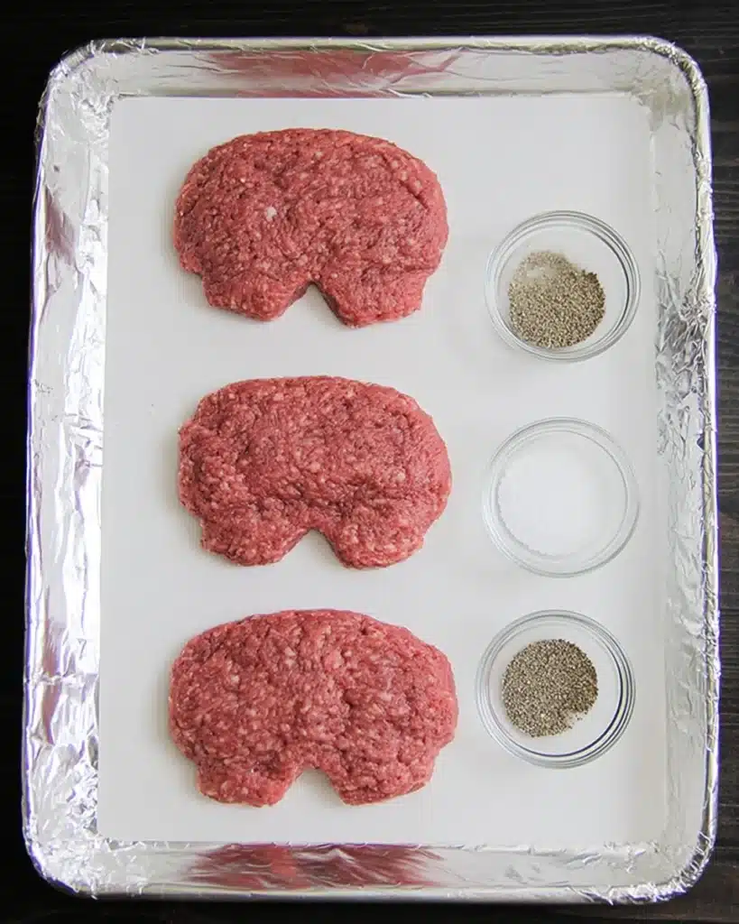 Raw ground beef patties shaped like skulls on a foil-lined baking tray with salt and pepper