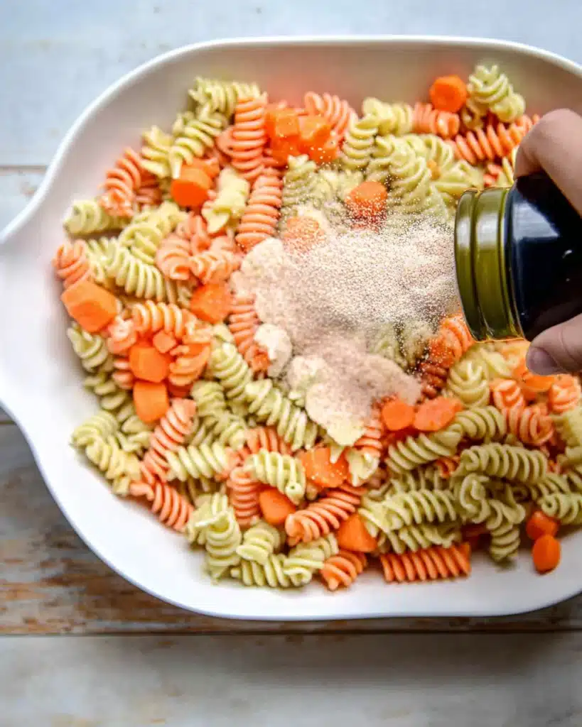 Halloween Pasta Salad – A Fun and Spooky Side Dish 12 “Tricolor pasta and carrots in a white bowl being sprinkled with garlic powder while preparing Halloween Pasta Salad.”