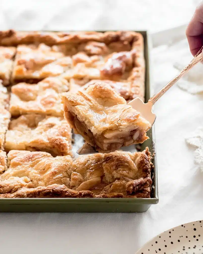 Freshly baked apple slab pie slice being lifted from pan, showing flaky crust and juicy apple filling.