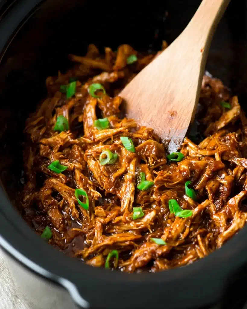 Shredded honey garlic chicken in a slow cooker with glossy sauce and green onions.