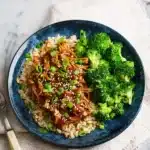 Shredded honey garlic chicken over brown rice with steamed broccoli, sesame seeds, and green onions on a blue plate.