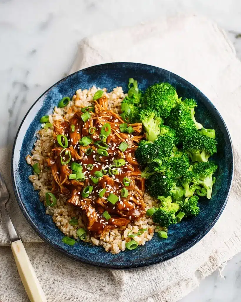 Shredded honey garlic chicken over brown rice with steamed broccoli, sesame seeds, and green onions on a blue plate.