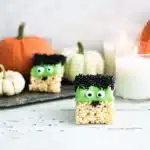 Halloween Rice Krispie treat decorated like a green monster with candy eyes, black sprinkle hair, and a crispy marshmallow base, styled with pumpkins and candles.