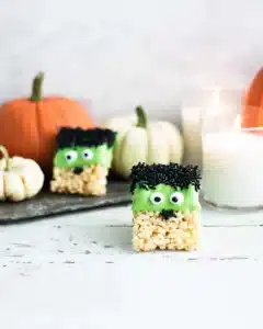 Halloween Rice Krispie treat decorated like a green monster with candy eyes, black sprinkle hair, and a crispy marshmallow base, styled with pumpkins and candles.