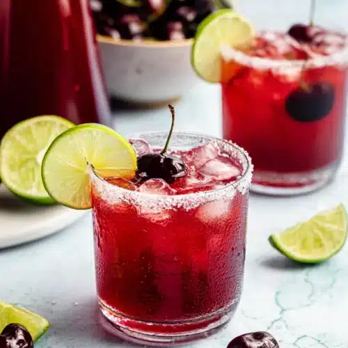 Spiked Cherry Limeade cocktails with fresh cherries, lime wedges, ice, and sugared rims